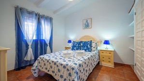 Une chambre à coucher avec un lit, deux tables de chevet avec des lampes, une fenêtre avec des rideaux bleus et un tableau accroché au mur.