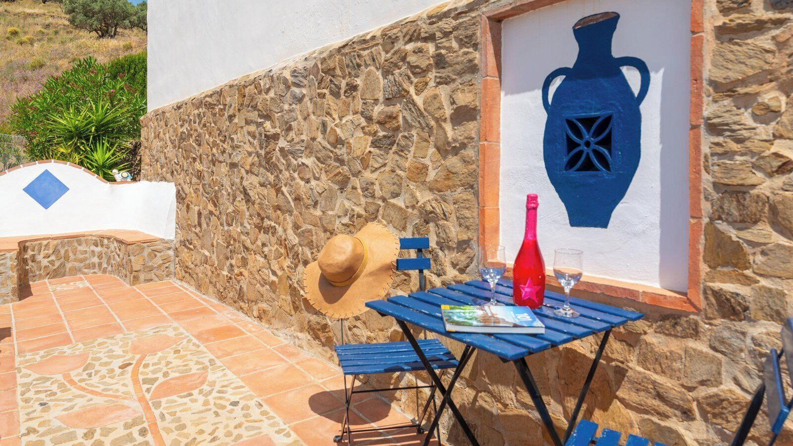 Un mur de pierres avec une terrasse carrelée, une table bleue avec une bouteille rouge et des verres, un chapeau de paille et un tableau encadré représentant un vase bleu.