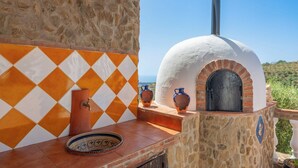 Un four en pierre doté d’une voûte blanche et d’une base en briques, un pare-feu en carreaux aux motifs losangés orange et blancs, et un comptoir en bois rustique agrémenté d’une assiette en céramique et de deux objets en poterie.