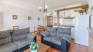 A living room with two sofas, a coffee table with flowers, a dining area with chairs, and a kitchen with a refrigerator and a hanging light fixture. - Casas Villanueva - Casa 2 Conil de la Frontera by Ruralidays (Cádiz)