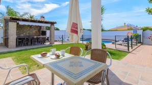 An outdoor patio with a table set for two, a stone barbecue area, and a pool in the background. - Casas Villanueva - Casa 2 Conil de la Frontera by Ruralidays (Cádiz)