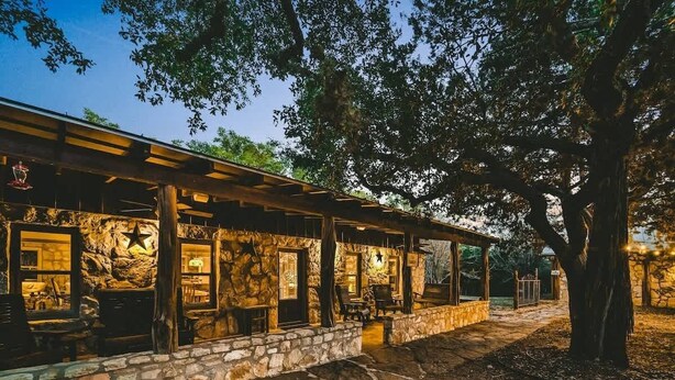 Un espacio de estar al aire libre de estilo rústico con muros de piedra y vigas de madera, que incluye mesas y sillas bajo un techo cubierto.