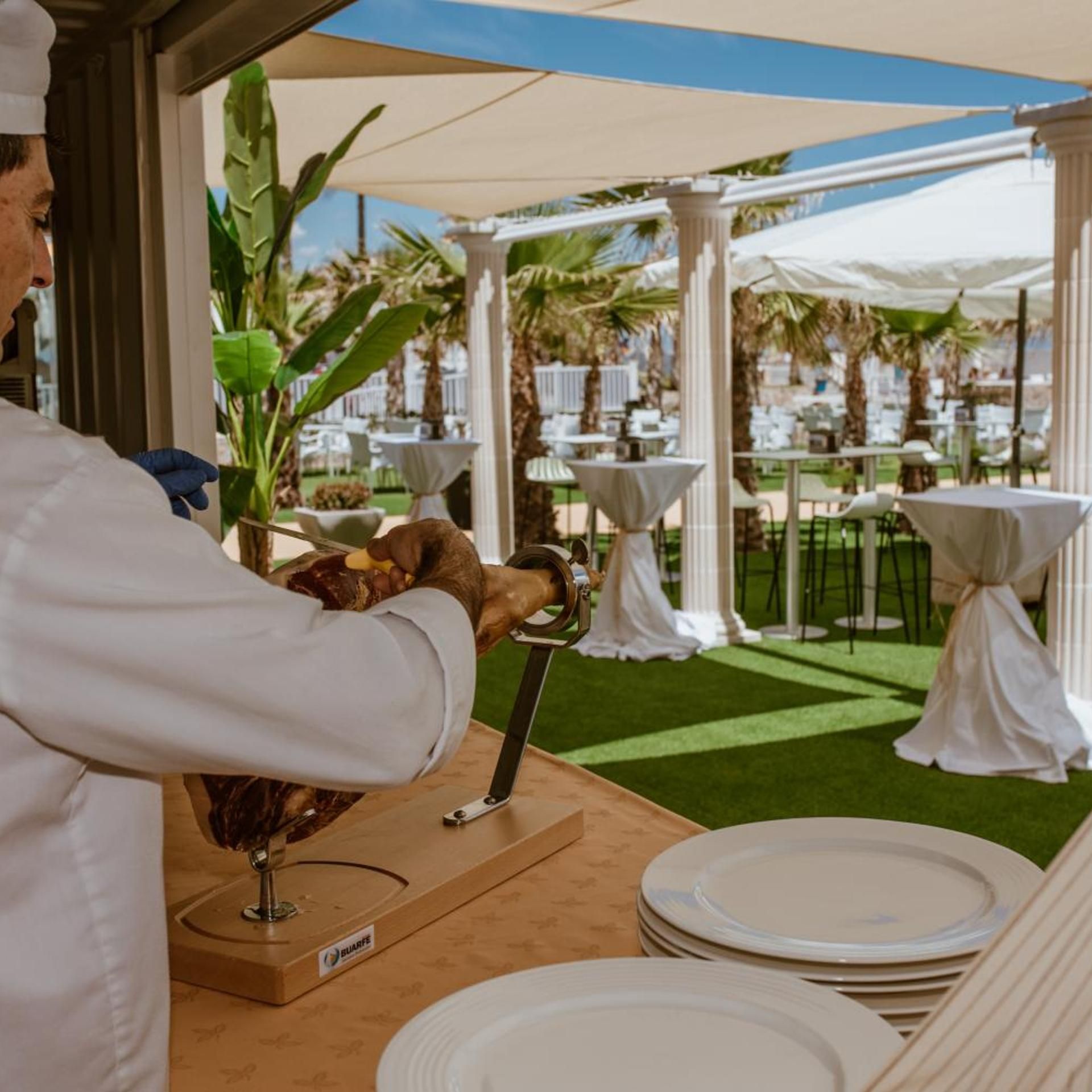 Un chef preparando comida en un área de comedor al aire libre con mesas blancas y columnas, rodeado de follaje.