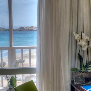 Quarto com uma grande janela com vista para uma praia e oceano, uma orquídea em vaso sobre uma mesa e uma cadeira.