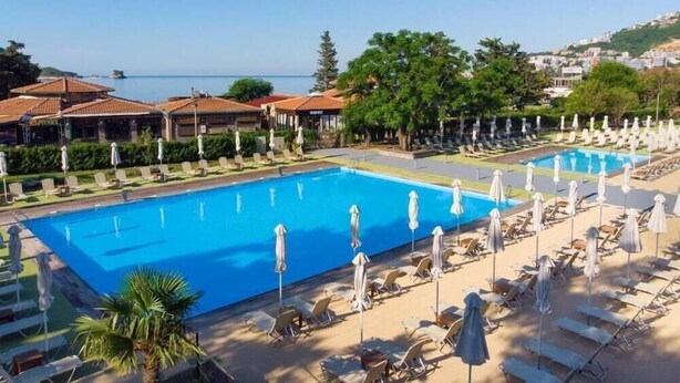 Zona de piscina con sillones de playa y sombrillas, vista despejada al mar y edificios con tejados rojos.