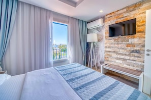 Habitación de hotel con una cama, un televisor montado en una pared de piedra, un equipo de aire acondicionado, una ventana con cortinas y vistas al exterior.