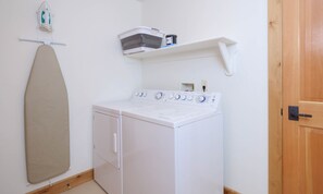 A laundry room with a washing machine, an ironing board, and a shelf with laundry supplies.