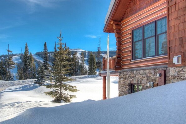 A log cabin with a stone foundation, large windows, and a view of a snowy mountain landscape.