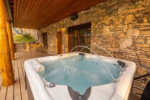 A hot tub on a wooden deck with stone walls and windows. - Platinum Big Sky Log Cabin | Ski-In/Out, Hot Tub, 2 King Suites & Arcade (Big Sky)