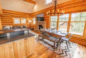 A log cabin interior with a stone fireplace, wooden dining table, and large windows. - Platinum Big Sky Log Cabin | Ski-In/Out, Hot Tub, 2 King Suites & Arcade (Big Sky)