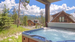 A hot tub with a wooden deck, surrounded by a forest and mountains in the background. - BIg Sky Vacation Rentals: PR Manitou 15 (Big Sky)