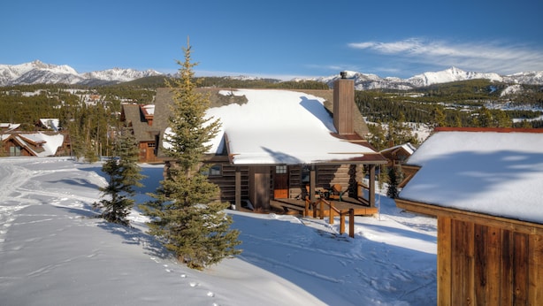 A log cabin with a snow-covered roof, surrounded by a snowy landscape and mountains in the background. - BIg Sky Vacation Rentals: PR Manitou 15 (Big Sky)