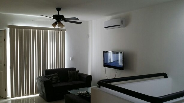A modern living room with a ceiling fan, a flat-screen TV mounted on the wall, a black sofa, and a glass coffee table.