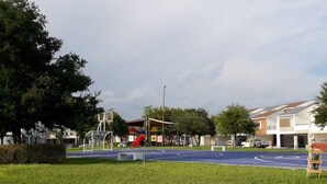 A playground with a blue surface, surrounded by grass and trees, with buildings in the background.