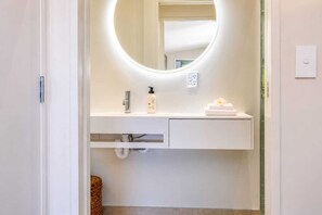 Een moderne badkamer met een ronde spiegel, een witte wastafel, een handdoek en een fles shampoo.