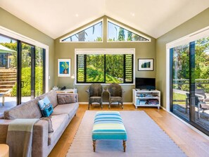 A spacious living room with a high ceiling, large windows, a sofa, chairs, a TV, and a bookshelf.