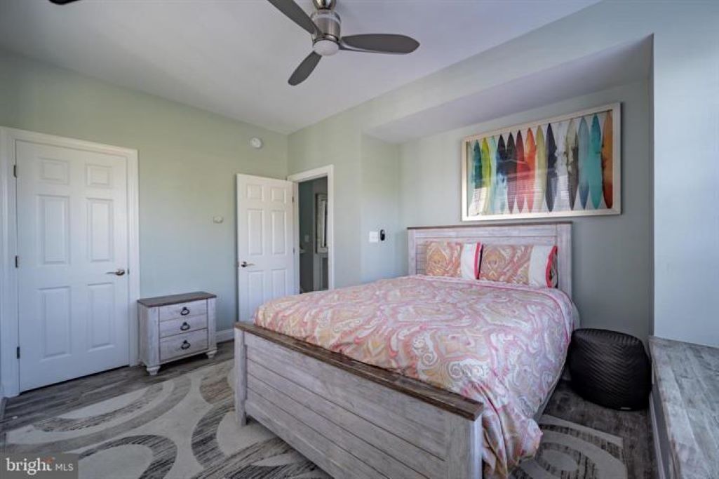A bedroom with a bed, a ceiling fan, a door, a dresser, and a colorful artwork on the wall.