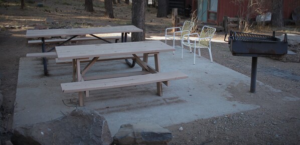 Un espace pique-nique avec des tables en bois et des chaises en métal, un barbecue et un bâtiment rouge en arrière-plan.