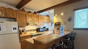 Una cocina con armarios de madera, una isla central con taburetes de bar, un refrigerador y una ventana con persianas.