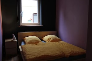 A small, dimly lit room with a single bed, a nightstand, and a window.