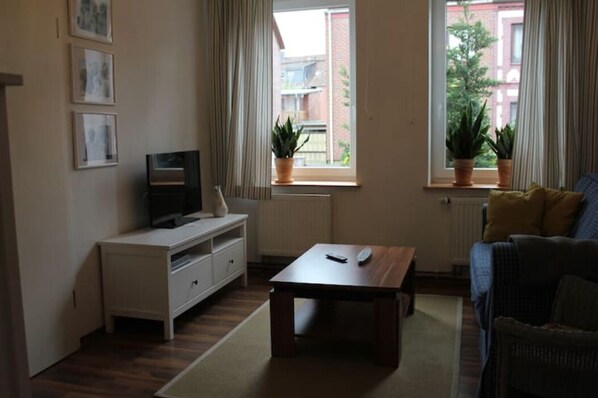A living room with a TV on a white cabinet, a wooden coffee table, a sofa, and two potted plants by the windows.