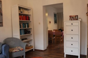 A room with a bookshelf, a chair, a white dresser, and a dining area in the background.