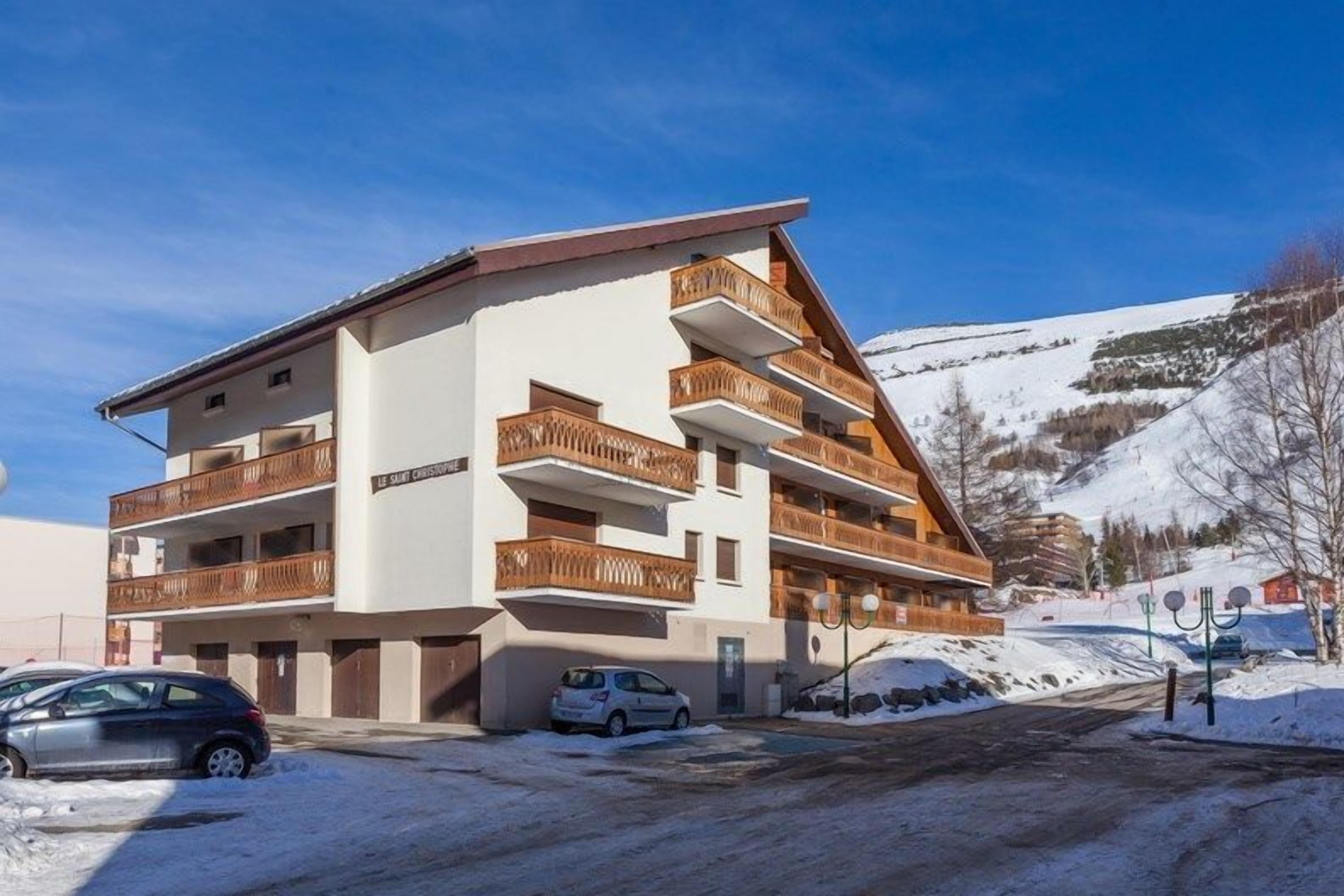 A multi-story hotel with balconies, a ski slope in the background, and parked cars.