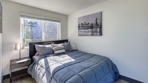 A bedroom with a bed, a nightstand, a lamp, a window with blinds, and a cityscape painting on the wall. - ► Modern Tiny House ◄ Big Personality, AC & Views! (Tukwila)