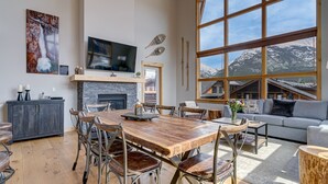 A spacious living area with a large wooden dining table, comfortable seating, a fireplace, a TV mounted on the wall, and a large window offering a mountain view. - Spectacular 3 bedroom Penthouse - Vaulted! Views! (Canmore)