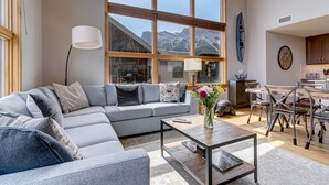 A modern living room with a grey sectional sofa, a coffee table with a vase of flowers, and a view of mountains through large windows. - Spectacular 3 bedroom Penthouse - Vaulted! Views! (Canmore)