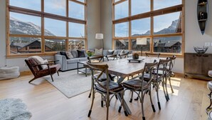 A spacious living area with large windows, a dining table, and a cozy seating arrangement. - Spectacular 3 bedroom Penthouse - Vaulted! Views! (Canmore)