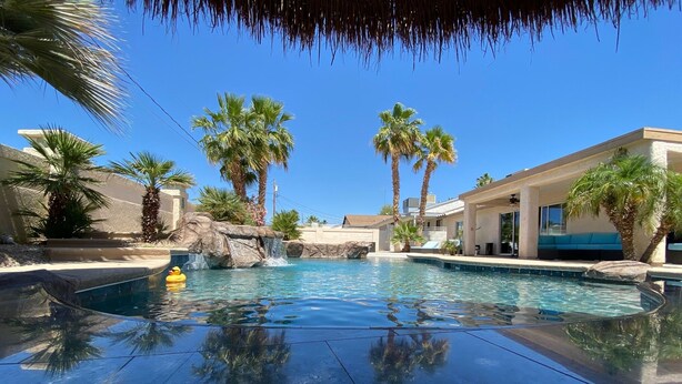 A swimming pool with a waterfall feature, surrounded by palm trees and modern buildings. - River Oasis (Lake Havasu City)