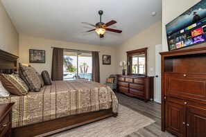 A bedroom with a large bed, a ceiling fan, a TV mounted on the wall, and a view of the outdoors.
