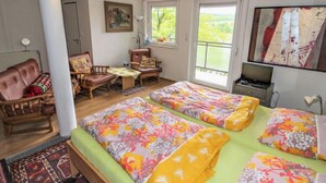 A room with two beds, a sofa, a chair, a table, and a TV. There is a window with a view of greenery outside.