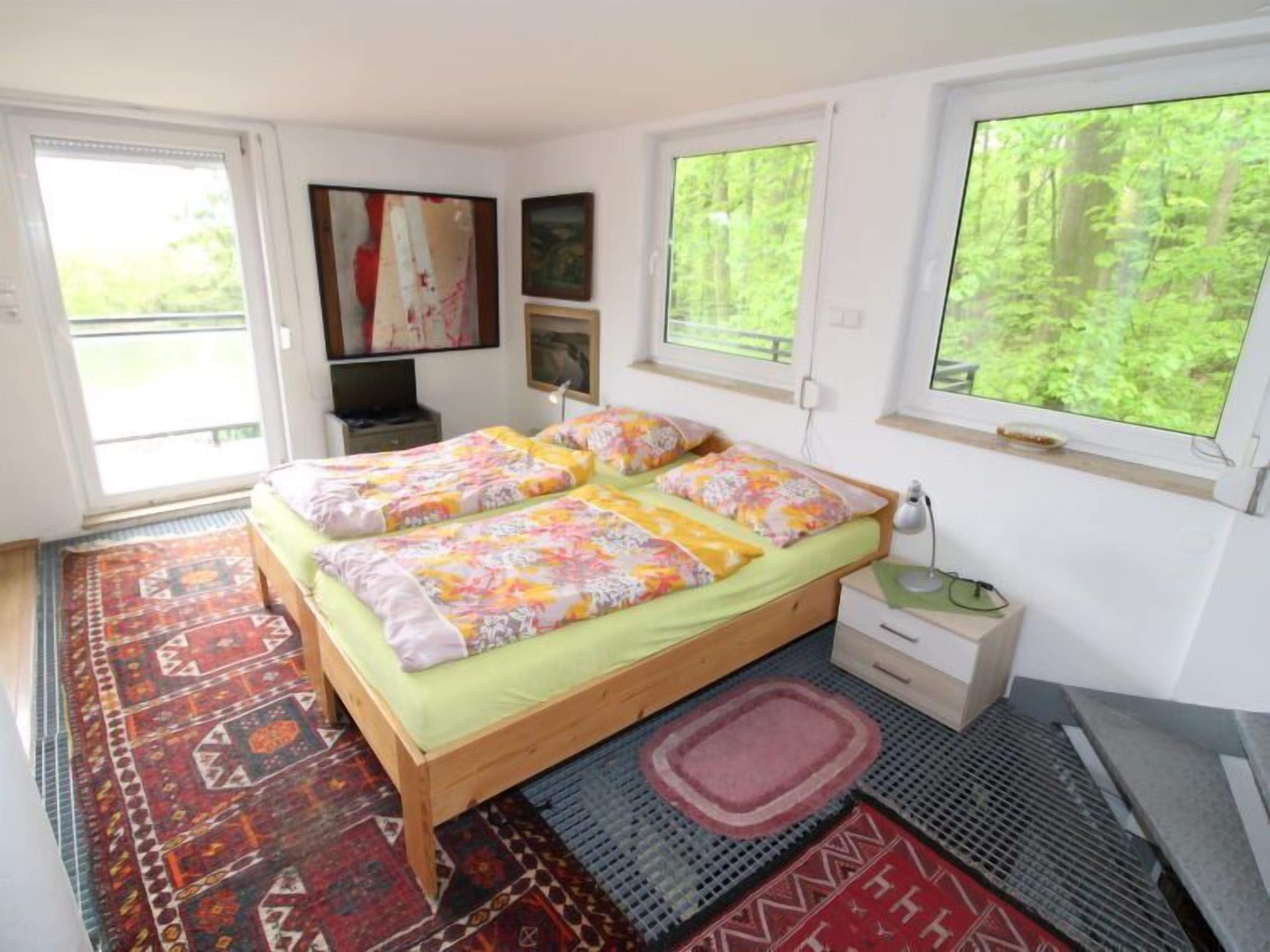 A bedroom with a bed, a nightstand, a desk, and a window with a view of greenery.