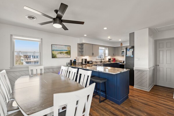 A modern kitchen with a central island, white chairs, a ceiling fan, and a view of buildings outside. - Beach House: 4-bedroom, Oceanfront, Awesome Views (Duck)