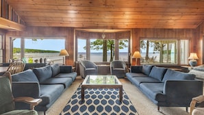 A cozy living room with a large window overlooking a beach, furnished with sofas, chairs, and a coffee table. - Higgins Lake Lakefront Lodge with Private Sandy Beach - Sleeps 13 (Roscommon)