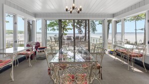 Une pièce spacieuse avec une grande table en verre, entourée de chaises et de tables en métal décorées, et de grandes fenêtres offrant une vue sur l’eau.