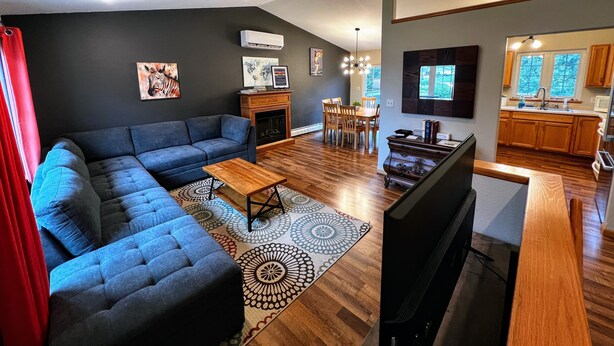 A modern living room with a sectional sofa, a wooden coffee table, and a TV. There is a dining area with a wooden table and chairs, and a kitchen with wooden cabinets and a window.