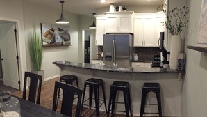 A modern kitchen with white cabinets, a stainless steel refrigerator, and a granite countertop. There are four black bar stools and a dining table with a glass jar on it. - Family-friendly Lakefront East Lake Okoboji Condo- Bridges Bay Resort (Arnolds Park)
