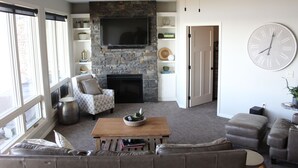 A living room with a stone fireplace, a flat-screen TV, a large clock, and a ceiling fan. - Family-friendly Lakefront East Lake Okoboji Condo- Bridges Bay Resort (Arnolds Park)