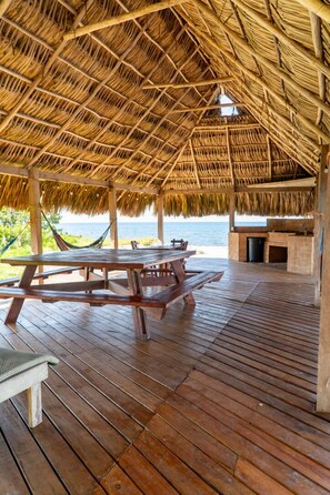Une terrasse en bois avec un toit de chaume, des tables de pique-nique et une vue sur l’océan.