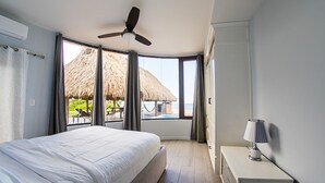 Une chambre avec un lit, un ventilateur de plafond, une table de chevet et une vue sur une plage avec une structure à toit de chaume.