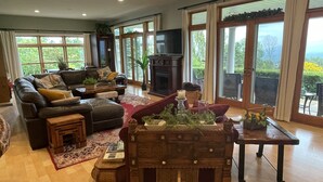 A spacious living room with a fireplace, leather sofa, and large windows offering a view of greenery. - Gentleman’s Farm Mountain Views Gourmet Kitchen Fire Pit Hike Ski Sleep 10 Pets! (Oak Hill)