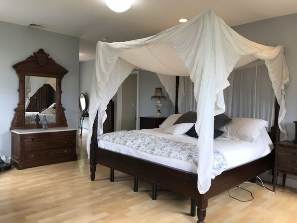 A four-poster canopy bed with a wooden frame and white bedding, a wooden dresser with a mirror, and a lamp on a bedside table.