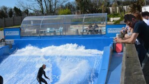 Piscina de olas con una persona practicando surf, rodeada de espectadores y una zona de estar cubierta.