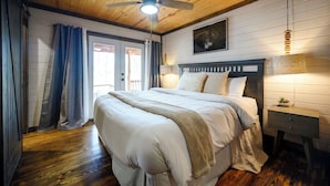 Quarto com teto de madeira, cama com roupa de cama branca, mesa de cabeceira, luminária na parede e ventilador de teto.