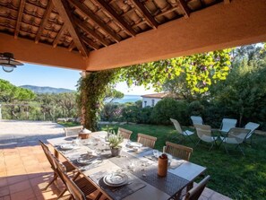 Ein Essbereich im Freien mit einem Tisch für sechs Personen, umgeben von Stühlen, unter einer Holzpergola mit Blick auf das Meer und die Hügel.