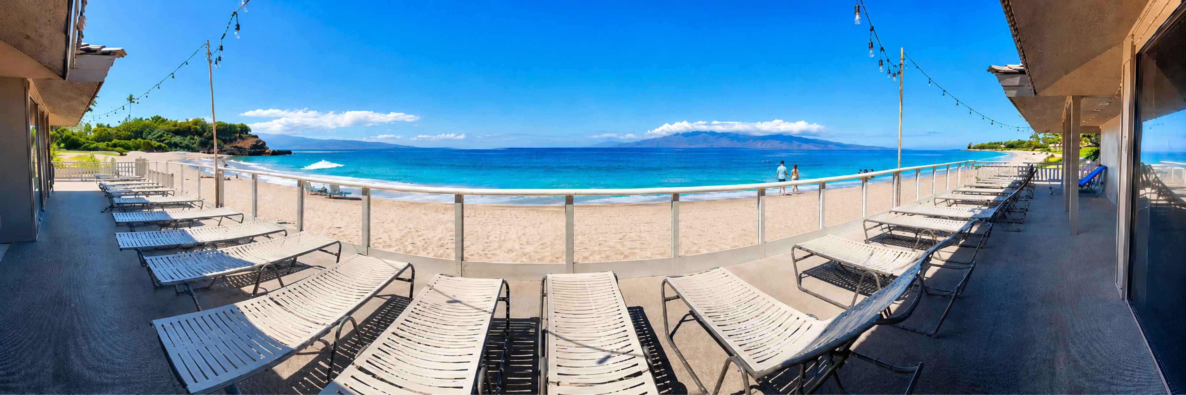 A beachside lounge with sun loungers facing the ocean, a clear sky, and a distant mountain range.