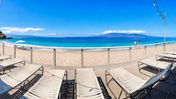A beachside lounge with sun loungers facing the ocean, a clear sky, and a distant mountain range.